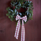 Ruby Cotton Christmas Bow in red check fabric with rounded loops and long tails, tied to a pine and eucalyptus wreath on a dark wood panelled door.