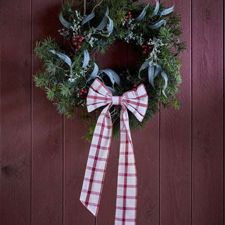 Ruby Cotton Christmas Bow in red check fabric with rounded loops and long tails, tied to a pine and eucalyptus wreath on a dark wood panelled door.