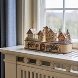 Norwich Wooden Christmas Decoration with Light in beige, a miniature wooden village with gabled houses, fir trees and figures, displayed on a windowsill.