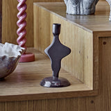 Cidra Aluminium Candle Holder in glossy brown, abstract curved silhouette with domed base, shown on a wooden shelf beside a pink twisted candlestick and ceramic bowl.