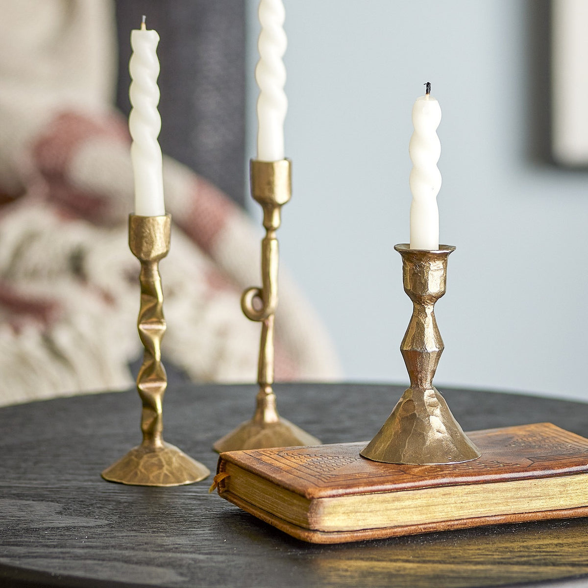 Efsa Iron Candle Holders Set of 3 in brass with hammered, faceted stems and bell-like bases, shown with white twisted candles on a dark table beside a leather-bound book.