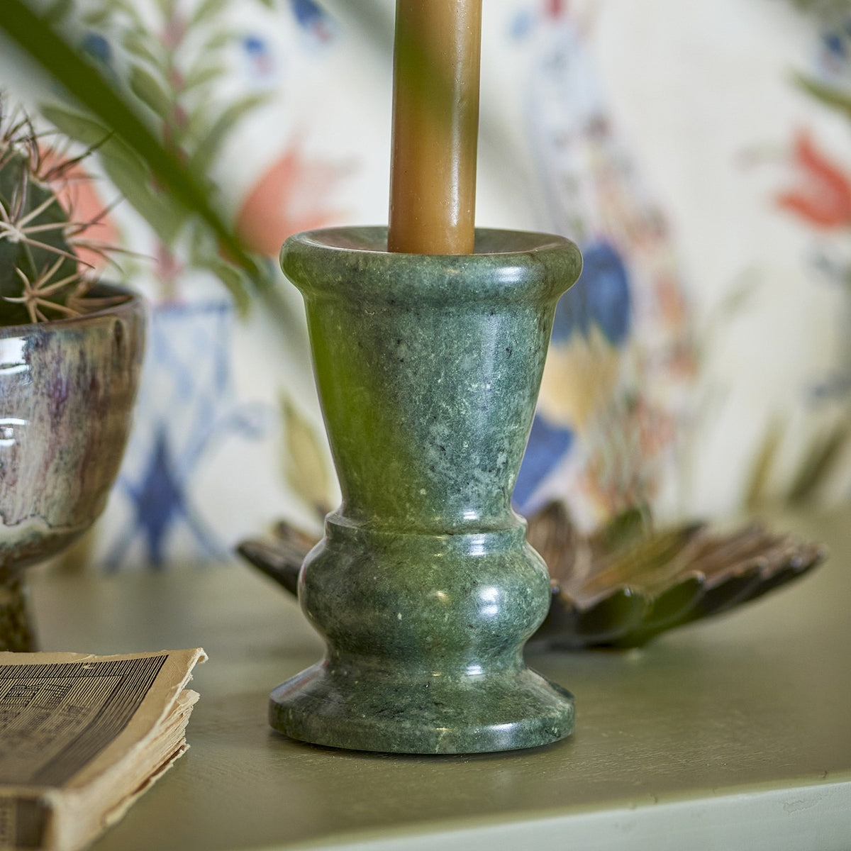 Sicilia Marble Candle Holder 12.5cm in green stone with pale veining, featuring a flared cup, narrow waist and stepped foot, holding a lit taper candle on a shelf.