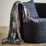 True Checked Cotton Throw in Multi, a woven cotton check blanket with thick shaggy fringe, draped over a dark upholstered armchair in a living room.