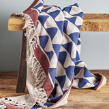 Rimme Cotton Throw in multicoloured navy and cream wave pattern with rust border and fringed edge, draped over a wooden bench.