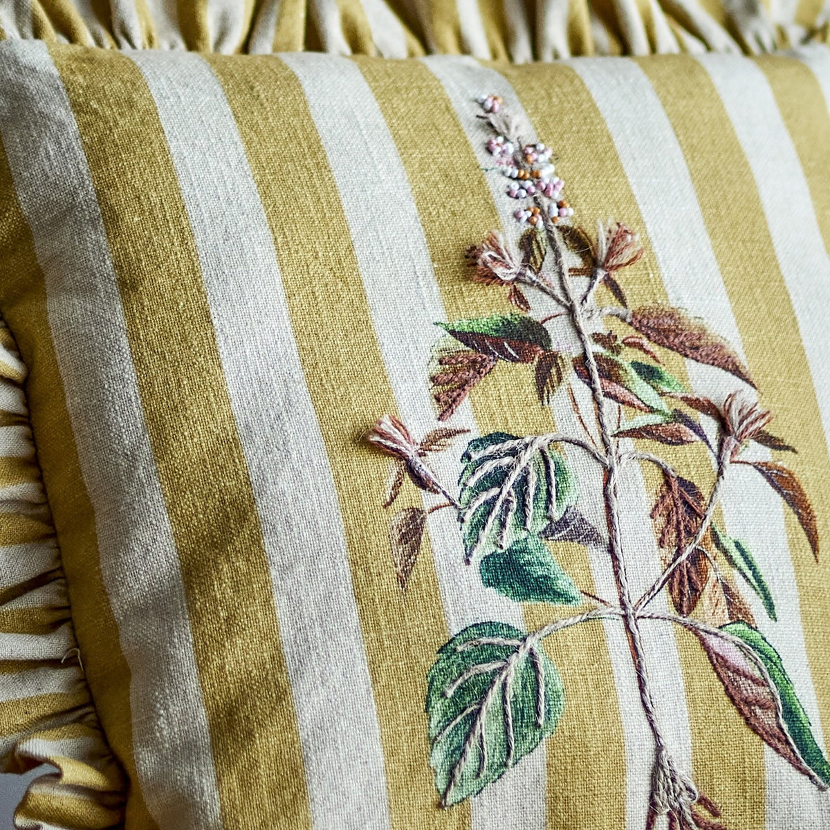 Camila Striped Cotton Cushion in yellow, close-up of woven stripes with an embroidered botanical stem and leaves, edged with a gathered ruffle in matching fabric.