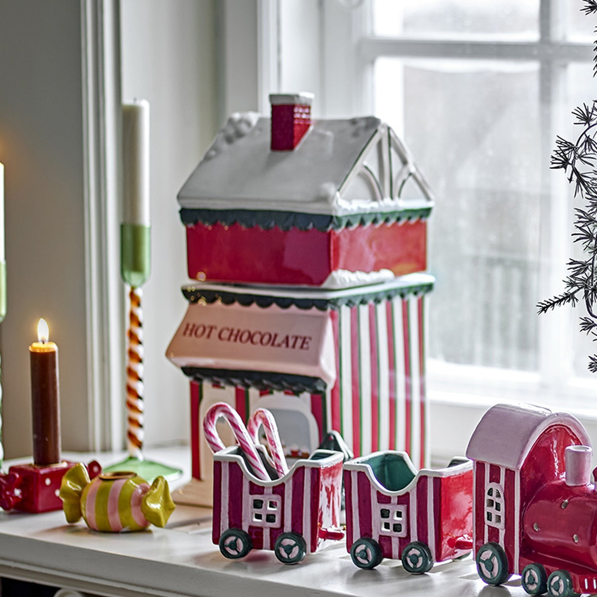 Gloria Ceramic Christmas Storage Jar in red, shaped like a shop with snow-topped roof, chimney and "HOT CHOCOLATE" awning, shown on a windowsill with festive ceramic train and candles.