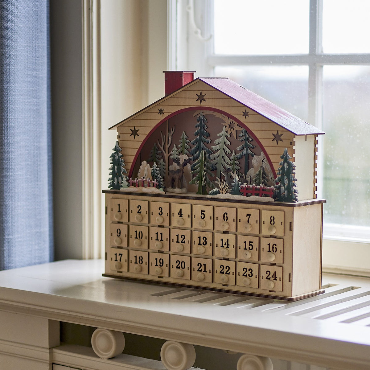 Melvin House Wooden Advent Calendar in beige plywood, with pitched roof, chimney, cut-out stars, arched winter forest scene and 24 numbered drawers, shown on a windowsill.