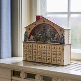 Melvin House Wooden Advent Calendar in beige plywood, with pitched roof, chimney, cut-out stars, arched winter forest scene and 24 numbered drawers, shown on a windowsill.
