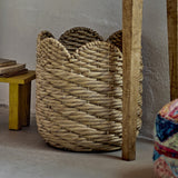Liam Water Hyacinth Basket in natural tone with a scalloped rim and diagonal woven pattern, shown beside a wooden bench and table leg in a living room setting.