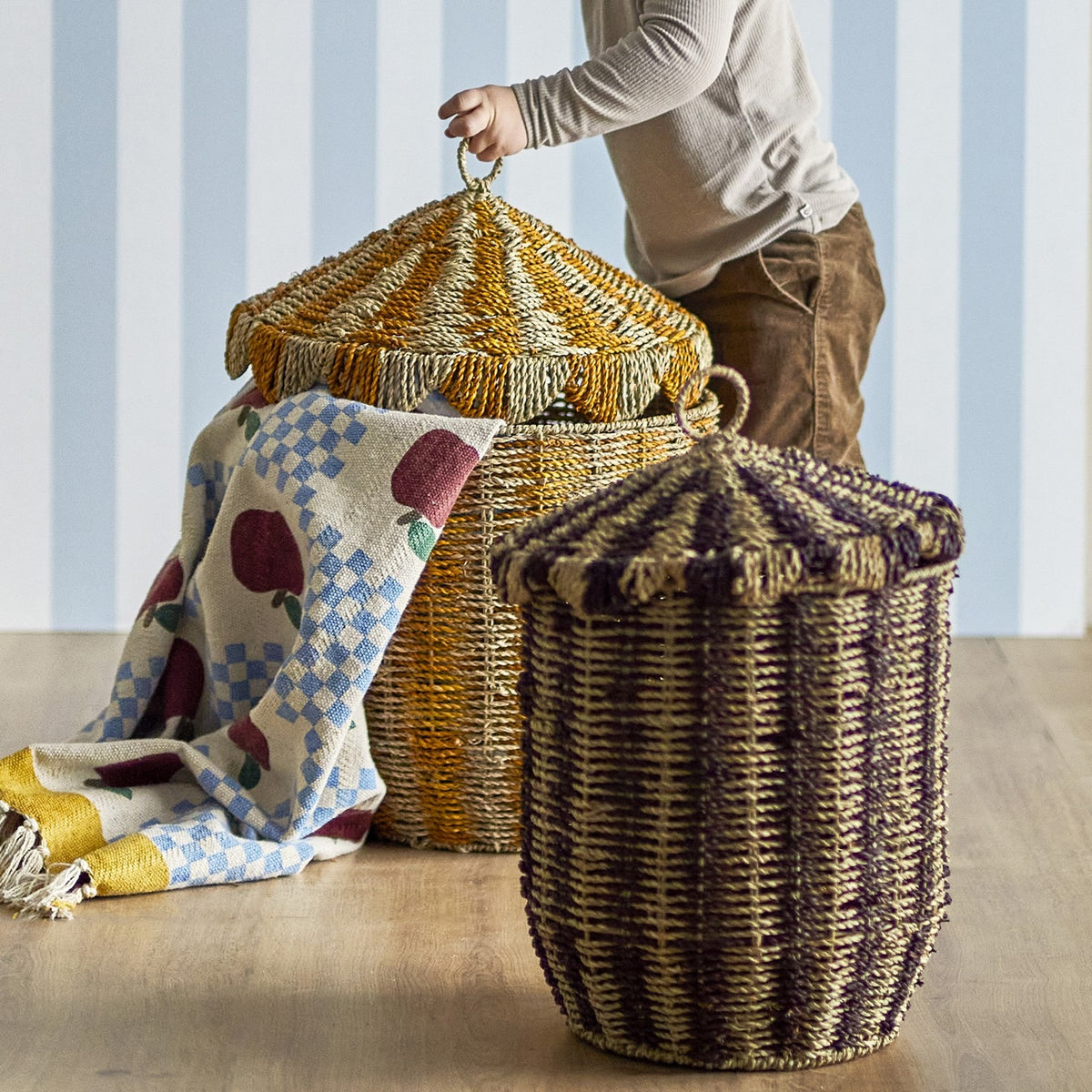 Cikkeline striped seagrass storage baskets with pointed lids and woven loop handles, shown in a nursery setting with a child and patterned blanket.