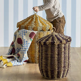Cikkeline striped seagrass storage baskets with pointed lids and woven loop handles, shown in a nursery setting with a child and patterned blanket.