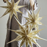 Ilo Paper Christmas Stars Set of 3 in cream, multi-pointed spiked shapes with cut-out detailing, shown hanging against a grey backdrop held by a person in a linen apron.