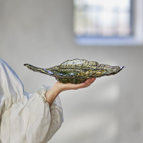 Aiku Glass Bowl 38x29cm in green, a wide shallow dish with rippled, textured rings, shown held in one hand against a blurred neutral interior backdrop.