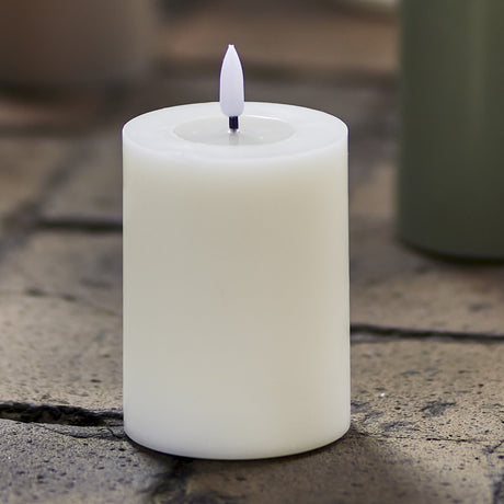 Ponza LED Candle 10cm in White, a flameless paraffin pillar with a realistic flame-tip and recessed top, shown outdoors on stone paving.