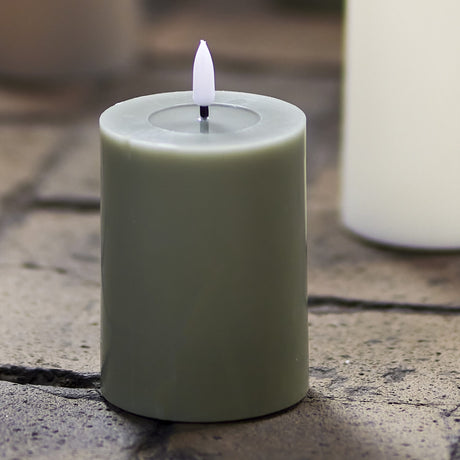 Ponza LED Candle 10cm in Green, a flameless pillar candle with paraffin-look wax finish and a white flame-shaped tip, shown outdoors on stone paving.