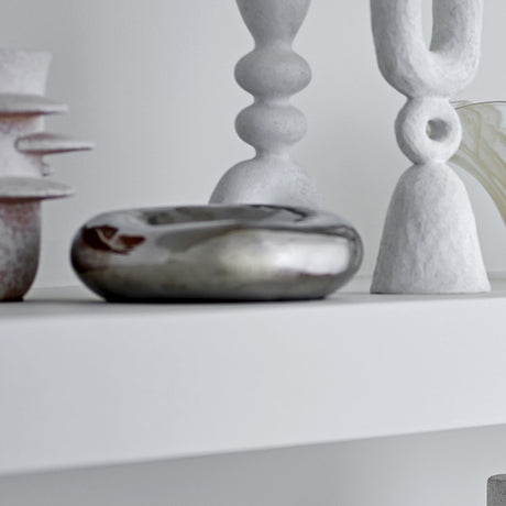 Shine Pebble Stoneware Bowl 18cm in silver, a low pebble-shaped dish with a mirror-like finish, shown on a shelf beside sculptural candlesticks and vases.