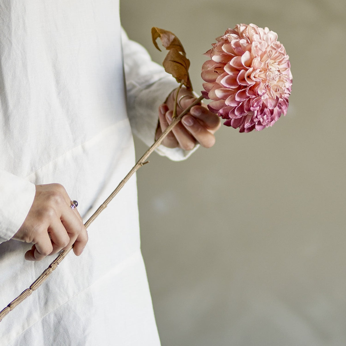 Hand holding the Chrysanthemum Artificial Stem 73cm in pink, showing the full rounded petal head and slender leafy stem against a neutral background.