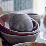 Paula Stoneware Cup 275ml in dark brown with a rounded body, loop handle, and speckled reactive glaze, nestled inside stacked ceramic bowls.