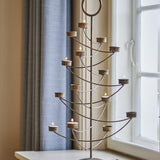 Alani Iron Christmas Tealight Holder in gold, a tree-shaped candle holder with a central stem, curved arms holding lit tealights, and a ring top, shown by a window with curtains.