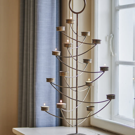 Alani Iron Christmas Tealight Holder in gold, a tree-shaped candle holder with a central stem, curved arms holding lit tealights, and a ring top, shown by a window with curtains.