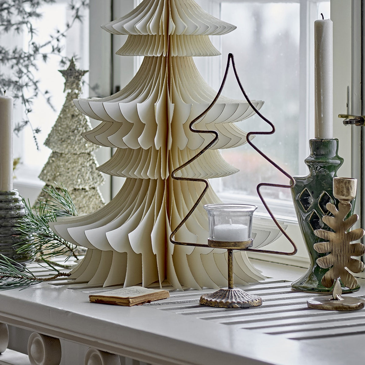 Miki Iron Christmas Tealight Holder in gold, a wire fir-tree outline with glass tealight cup, displayed on a windowsill among paper and wire tree decorations.