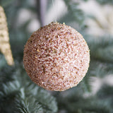 Nicolet Christmas Bauble in champagne, a round 9cm hanging decoration densely textured with beaded glitter, shown on a hanging thread against a blurred Christmas tree.