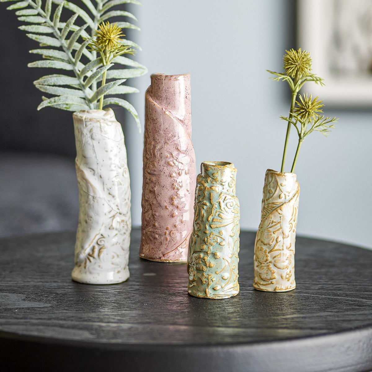 Set of four Temple Embossed stoneware vases in white, pink, green and cream with swirling textured patterns, holding foliage on a dark round table.