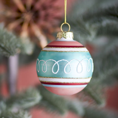 Kota Glass Christmas Bauble in multi colour, round glass ornament with pink, teal, white and red striped bands and a looping white pattern, gold hanging cap, pictured on a fir tree.