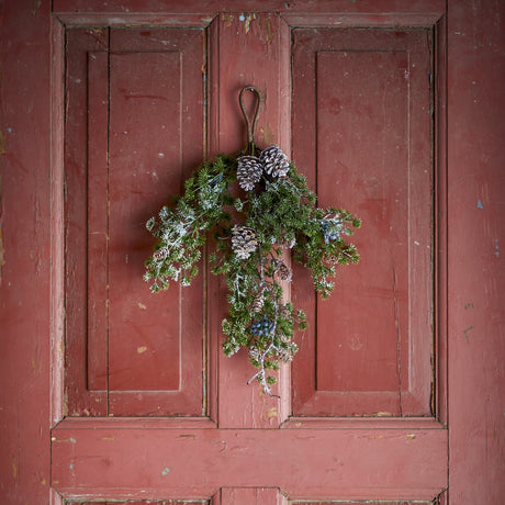 Cedar Christmas Decoration in green, a hanging faux spray with pinecones, berry clusters and frosted needle tips, hung by a loop on a weathered pink panelled door.