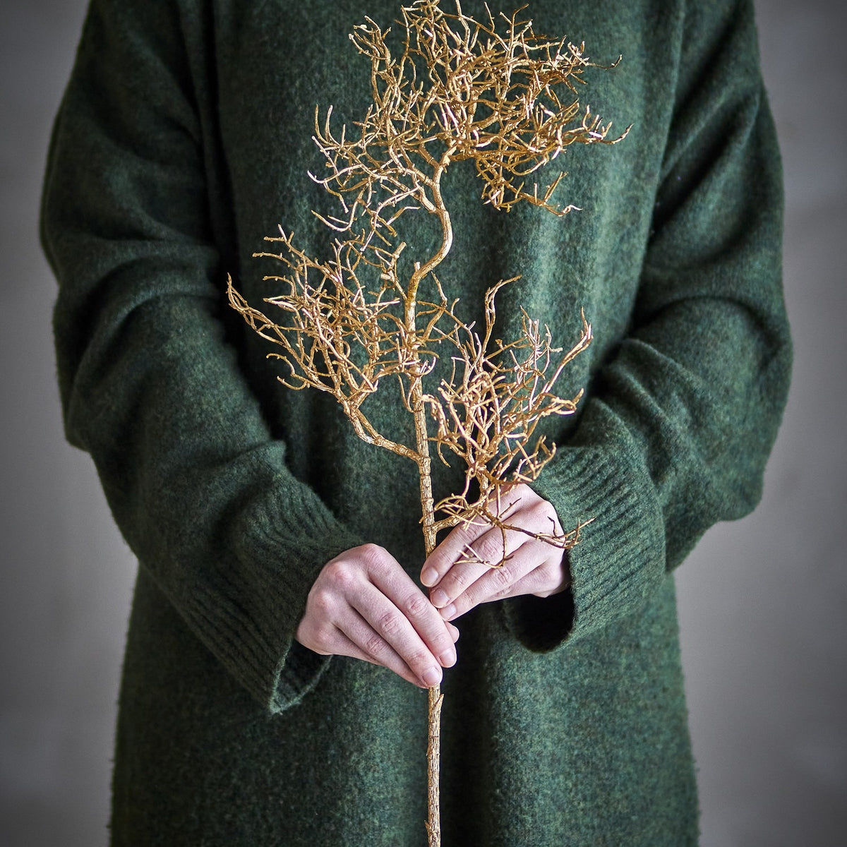 Person in a green jumper holding a gold artificial coral Christmas stem with a slim stalk and twiggy branching sprays, against a neutral grey background.