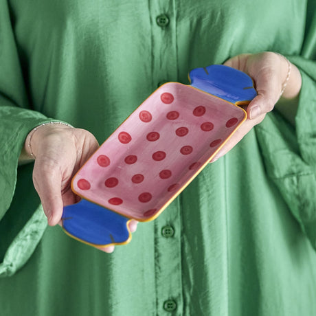 Hands hold the Caramella Wrapped Sweet stoneware platter, shaped like a twisted sweet wrapper in blue and yellow, with a pink interior decorated in hand-painted red polka dots.