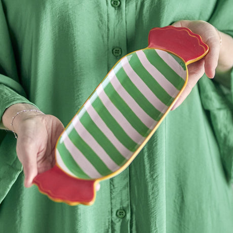 Caramella Striped Wrapped Sweet Christmas Platter in ceramic, with green and white diagonal stripes and red frilled wrapper ends, held in two hands against a green shirt.