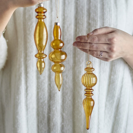 Auralia Glass Christmas Tree Decorations in gold, set of three slim finial-shaped drops with ribbed, fluted and spindle details, held by hand against a light fabric backdrop.