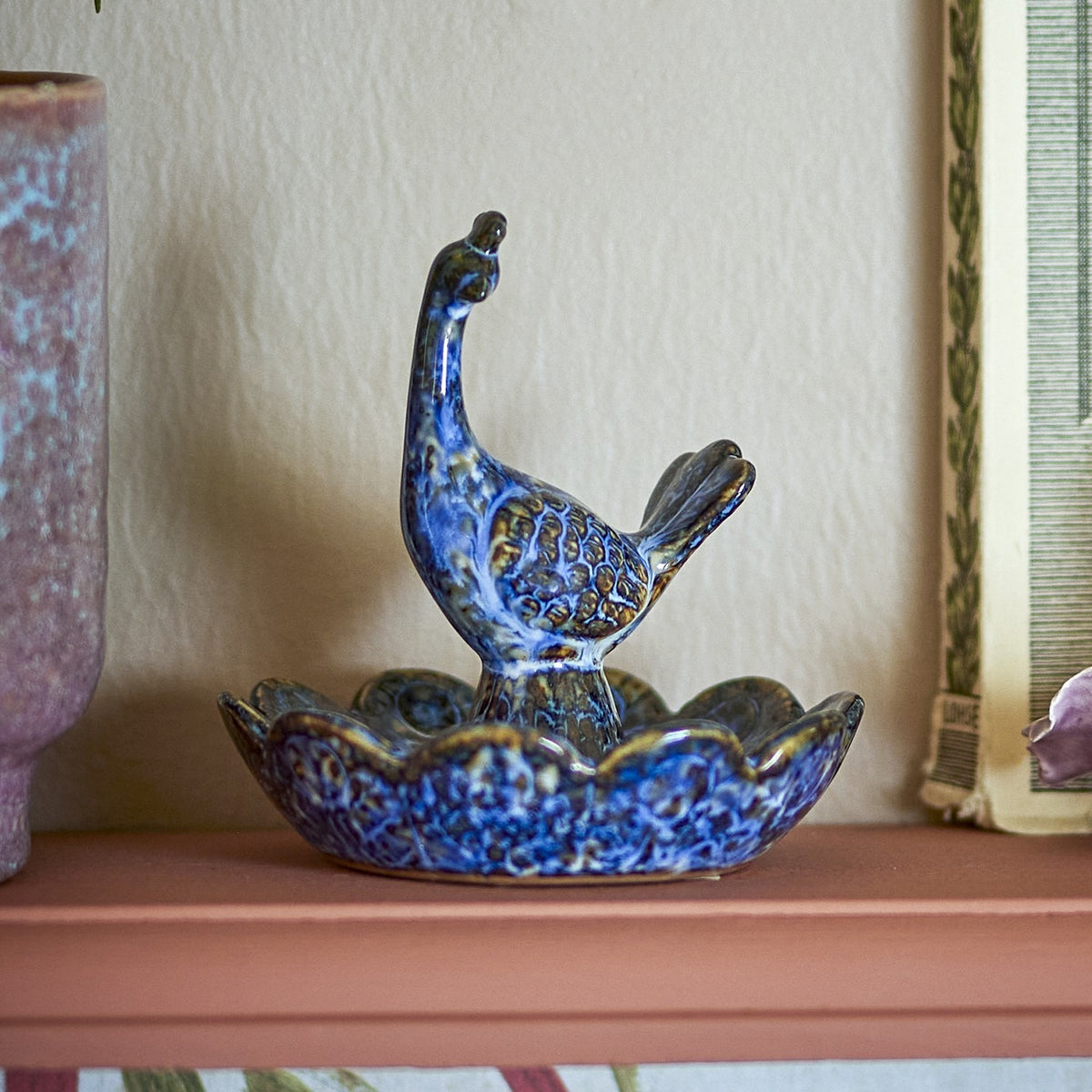 Kali Flower Stoneware Bowl 9.5cm in blue, a scalloped ceramic dish with a bird figure rising from a central pedestal, shown on a shelf beside a mirror and vase.
