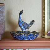 Kali Flower Stoneware Bowl 9.5cm in blue, a scalloped ceramic dish with a bird figure rising from a central pedestal, shown on a shelf beside a mirror and vase.