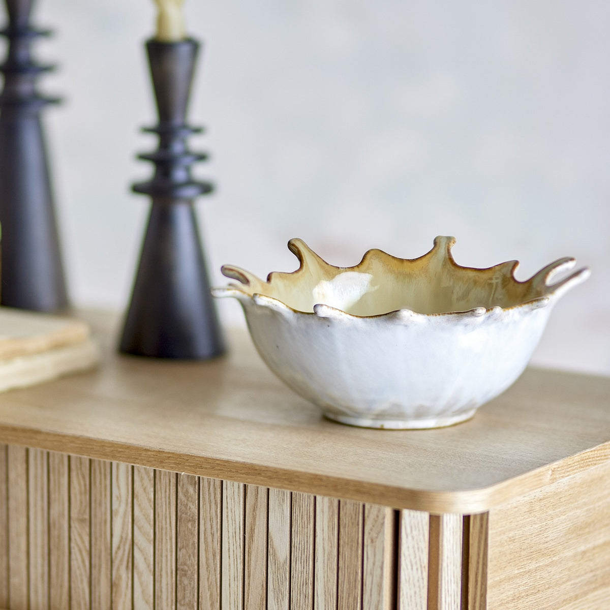 Alcott Stoneware Bowl 24x21cm in beige with a wavy, splash-edged rim and cream interior, shown on a wooden sideboard beside dark candlestick holders.