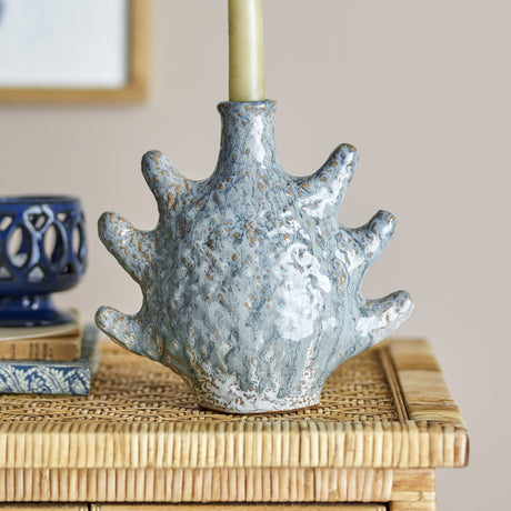 Bridget Shell Stoneware Candle Holder in grey, a fan-shaped ceramic base with six rounded arms and a narrow neck holding a taper candle, shown on a woven side table.