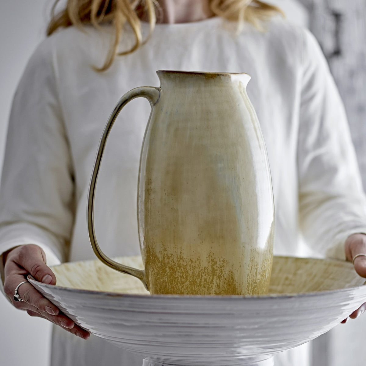 Saskia Speckled Stoneware Vase 31.5cm in beige, jug-shaped with looped handle and pinched rim, held atop a ridged white bowl by a person in a white dress.