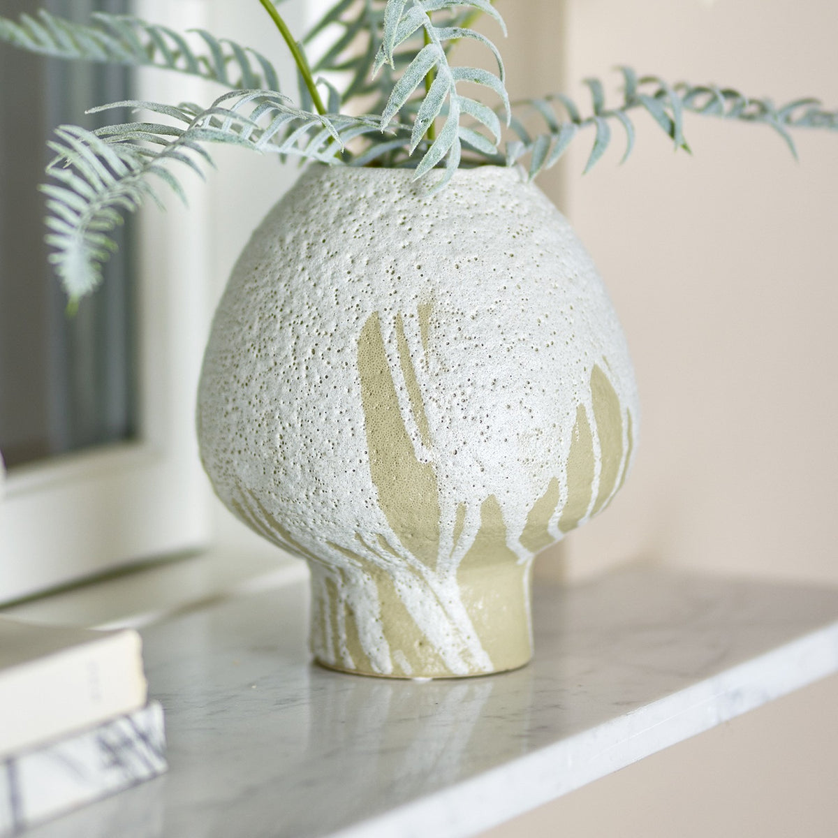 Morag Mushroom Stoneware Vase 19cm in White, with a rounded mushroom-shaped body in pitted, speckled glaze breaking into streaks near the foot, shown on a marble shelf with trailing foliage.