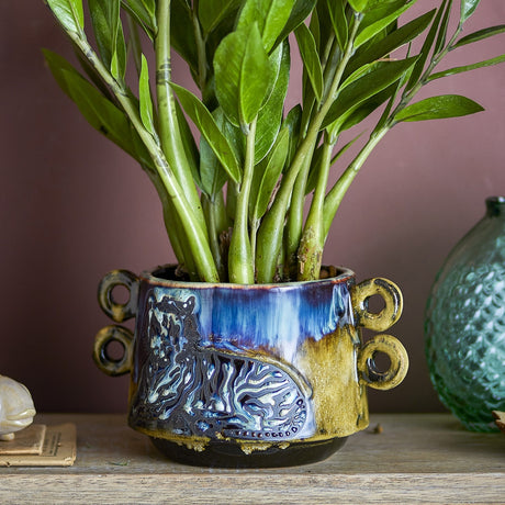 Walton Embossed Stoneware Plant Pot in multi glaze with raised tiger motif and stacked ring handles, shown holding a green leafy plant against a dusty pink wall.