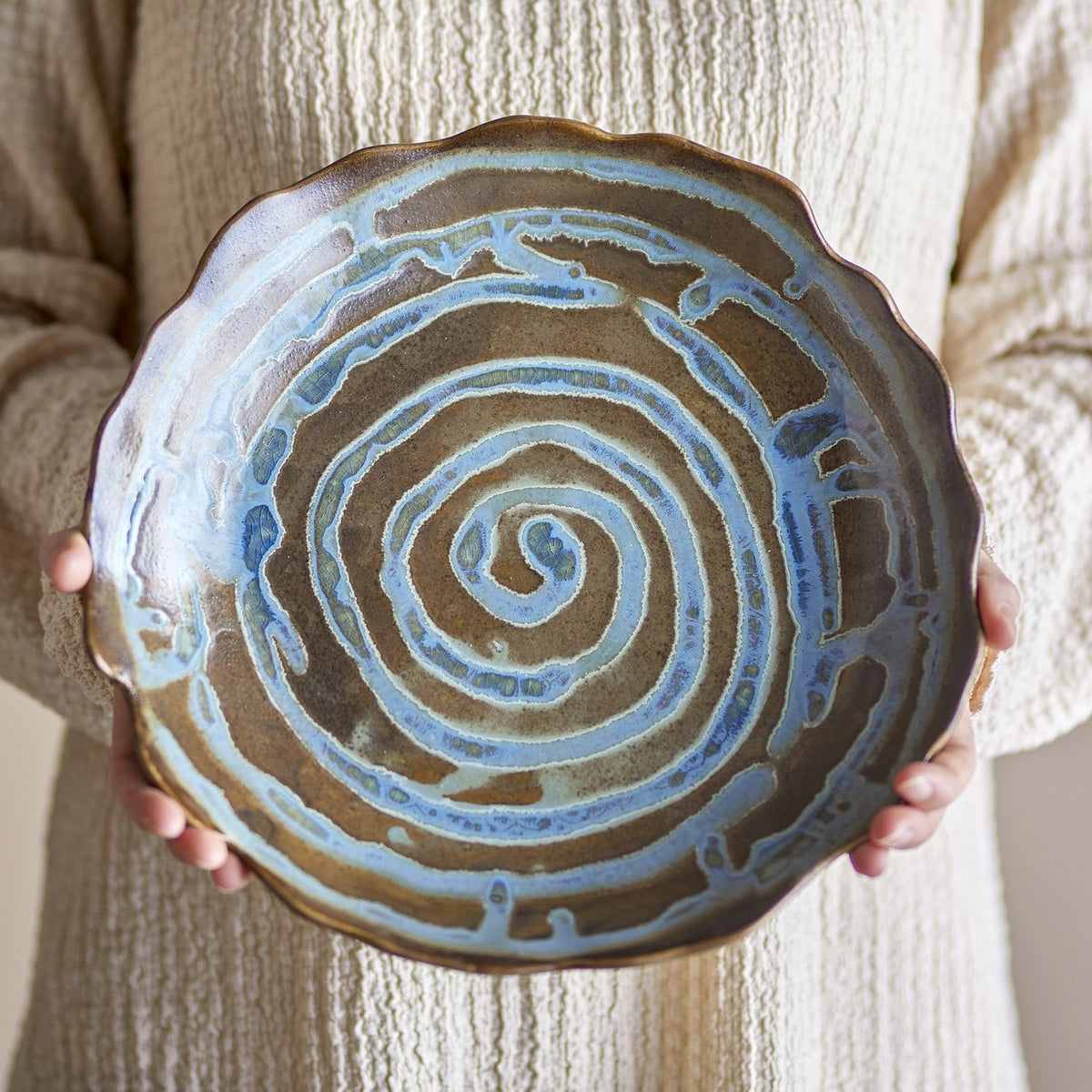 Marjorie Round Stoneware Tray in brown, held in both hands, showing a spiral glaze pattern and scalloped rim on a shallow ceramic serving dish.