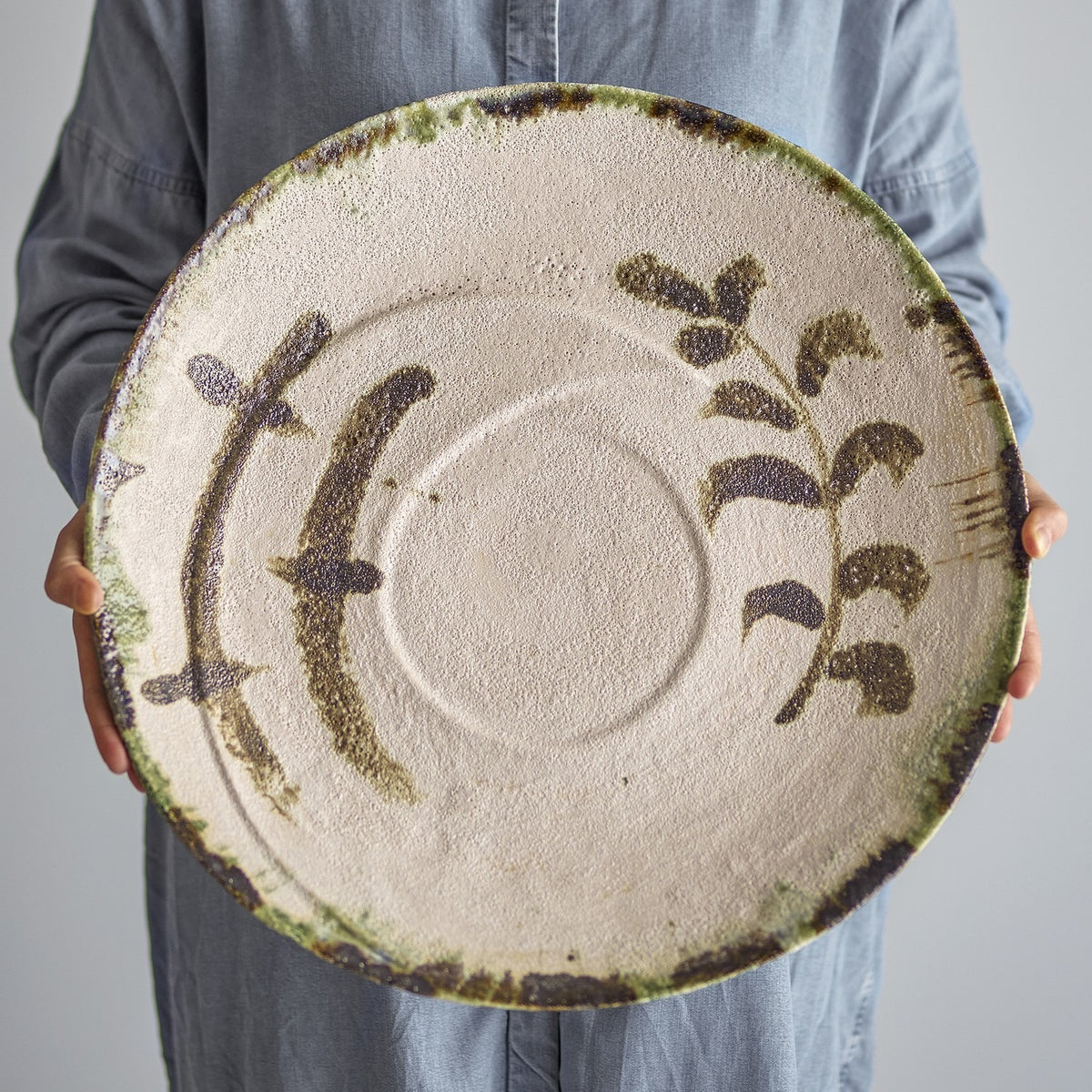 Hadyn Speckled Stoneware Tray in white, a large round tray with painted plant branches, a shallow central ring, and a dark speckled rim, held by a person in blue.