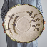 Hadyn Speckled Stoneware Tray in white, a large round tray with painted plant branches, a shallow central ring, and a dark speckled rim, held by a person in blue.
