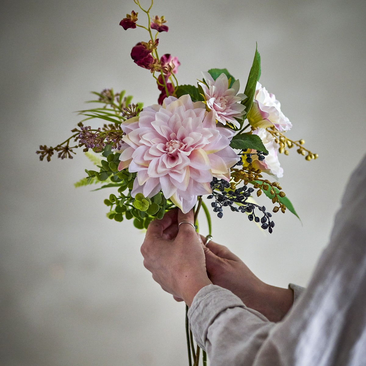 Splendor Artificial Bouquet in Pink held by hand, showing a large pink dahlia with ferns, berries and sprigs against a plain light background.