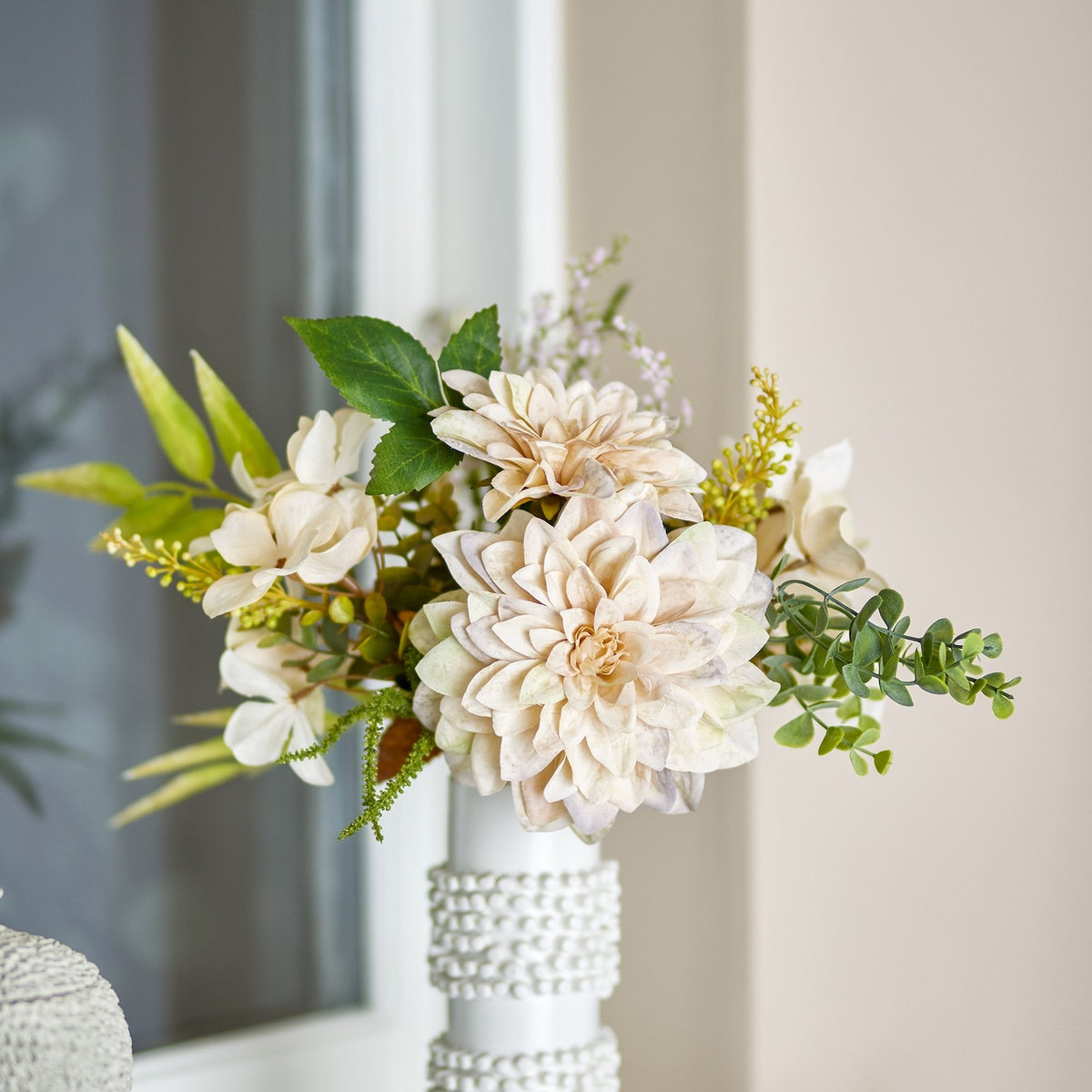Cream Glow artificial bouquet with dahlia blooms, hydrangea florets, fern fronds and eucalyptus sprigs in a white beaded vase, set in a living room.