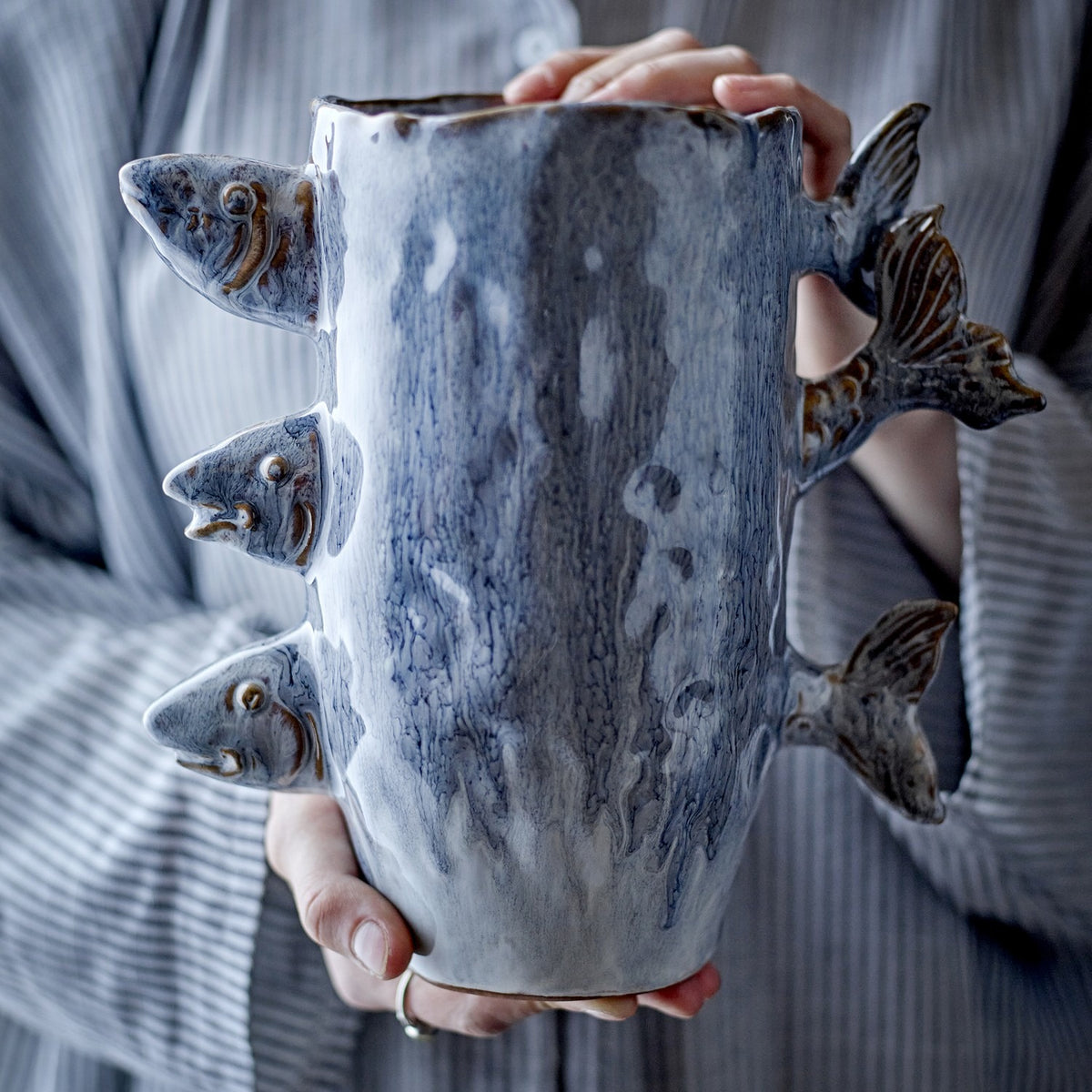 Tace Stoneware Vase 23cm in blue, with sculpted fish heads and tails protruding from the sides, held in hands against a striped grey garment.