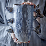 Tace Stoneware Vase 23cm in blue, with sculpted fish heads and tails protruding from the sides, held in hands against a striped grey garment.