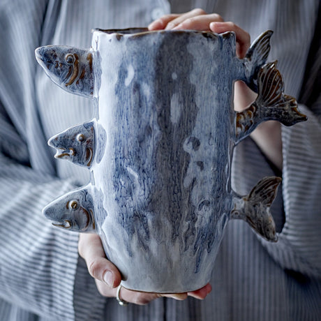 Tace Stoneware Vase 23cm in blue, with sculpted fish heads and tails protruding from the sides, held in hands against a striped grey garment.