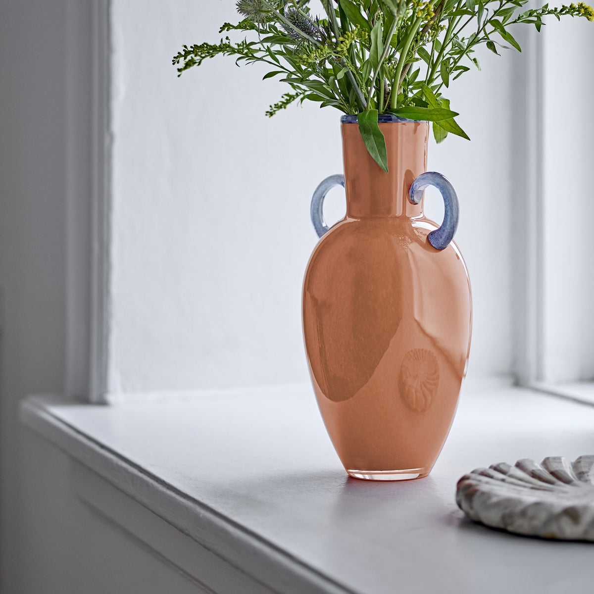 Anniversary Glass Vase 30cm in orange, amphora-shaped with blue loop handles and rim, holding greenery on a white windowsill.