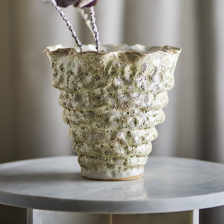 Colleen Wavy Stoneware Plant Pot in cream, round with a ruffled rim and stacked ridged body on a small foot, shown with foliage on a marble table against a neutral curtained backdrop.